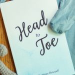 The Blue edition of the Head-to-Toe Prayer Journal. It is laying on a set of blue baby pajamas with a grey knitted teddy bear next to it.