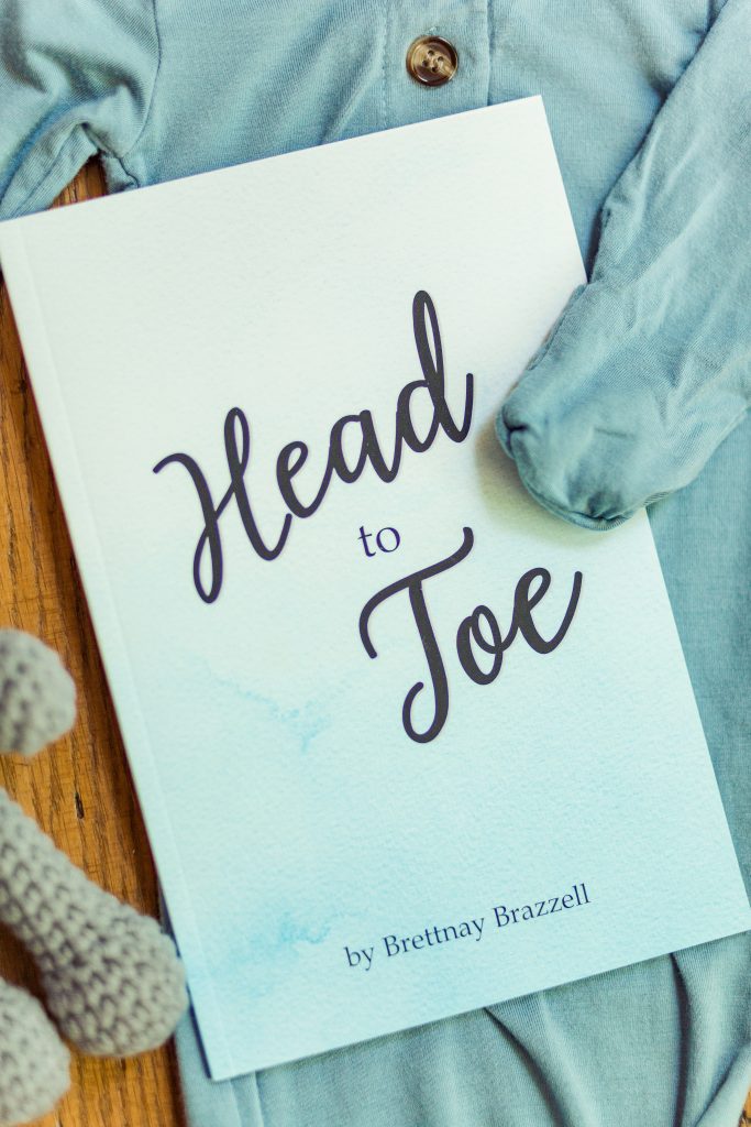 The Blue edition of the Head-to-Toe Prayer Journal. It is laying on a set of blue baby pajamas with a grey knitted teddy bear next to it.