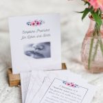 labor and delivery cards. It depicts the front card in a wooden stand with other cards flayed out in front of it. They are laying on a white blanket with a small vase of pink flowers beside them.