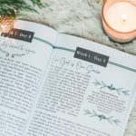 Great Hymns for Growing Hearts - Christmas Edition. The book is open to display the devotional content page. It is laying on a furry, white rug with a candle and evergreens around it.