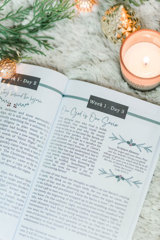 Great Hymns for Growing Hearts - Christmas Edition. The book is open to display the devotional content page. It is laying on a furry, white rug with a candle and evergreens around it.