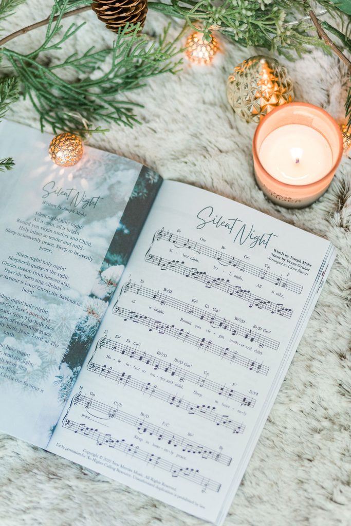 Great Hymns for Growing Hearts - Christmas Edition. The book is open to show the music to "Silent Night." It is laying on a furry, white rug with a candle and evergreens around it.