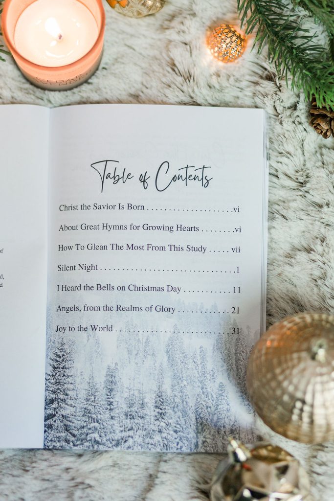 Great Hymns for Growing Hearts - Christmas Edition. The book is open to the table of contents. It is laying on a furry, white rug with a candle and evergreens around it.