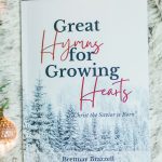 Great Hymns for Growing Hearts - Christmas Edition. The book has a snowy forest background. It is laying on a furry, white rug with a candle and evergreens around it.