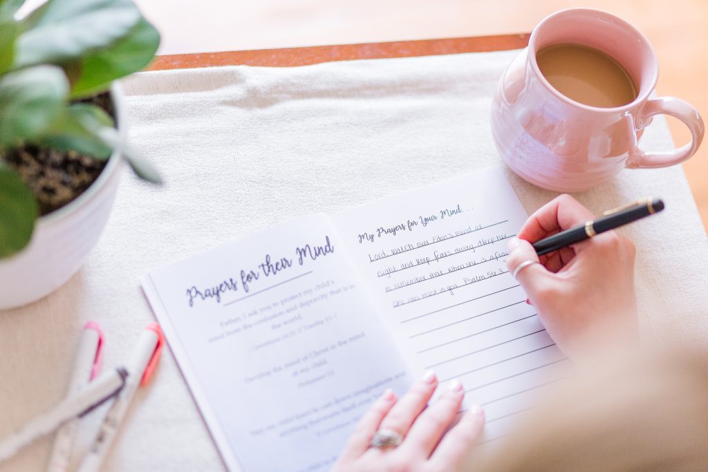 The Head to Toe Prayer Journal laying open on a table. A woman's hands are shown writing in it. There is a plant, pens, and a coffee next to the book.