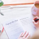 The Head to Toe Prayer Journal laying open on a table. A woman's hands are shown writing in it. There is a plant, pens, and a coffee next to the book.