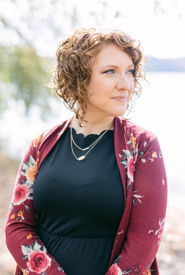 Casie Ravert is standing in front of a lake looking off to the right of the frame. She is wearing a red floral sweater over a black dress.