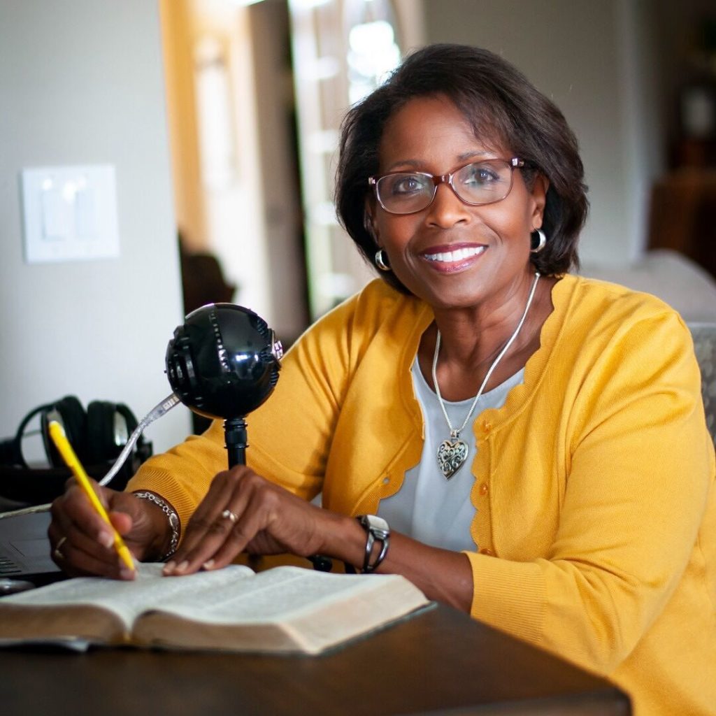 Francie Taylor is sitting at a desk with a Bible and microphone. She has a yellow pen in hand which matches her yellow top. She is smiling at the camera.