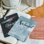 Three of the "Children of Character cards" laying on an open Bible which is on a wooden table. The center card reads "Anchored" with some descriptive text under it. The table also has a plant and a candle on it.
