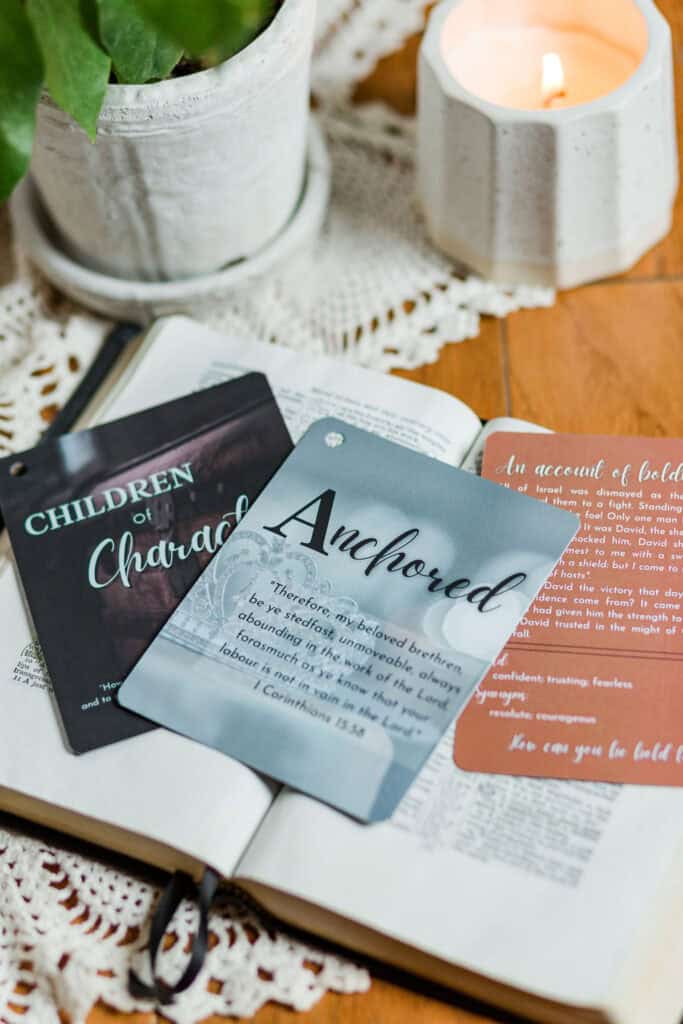 Three of the "Children of Character cards" laying on an open Bible which is on a wooden table. The center card reads "Anchored" with some descriptive text under it. The table also has a plant and a candle on it.