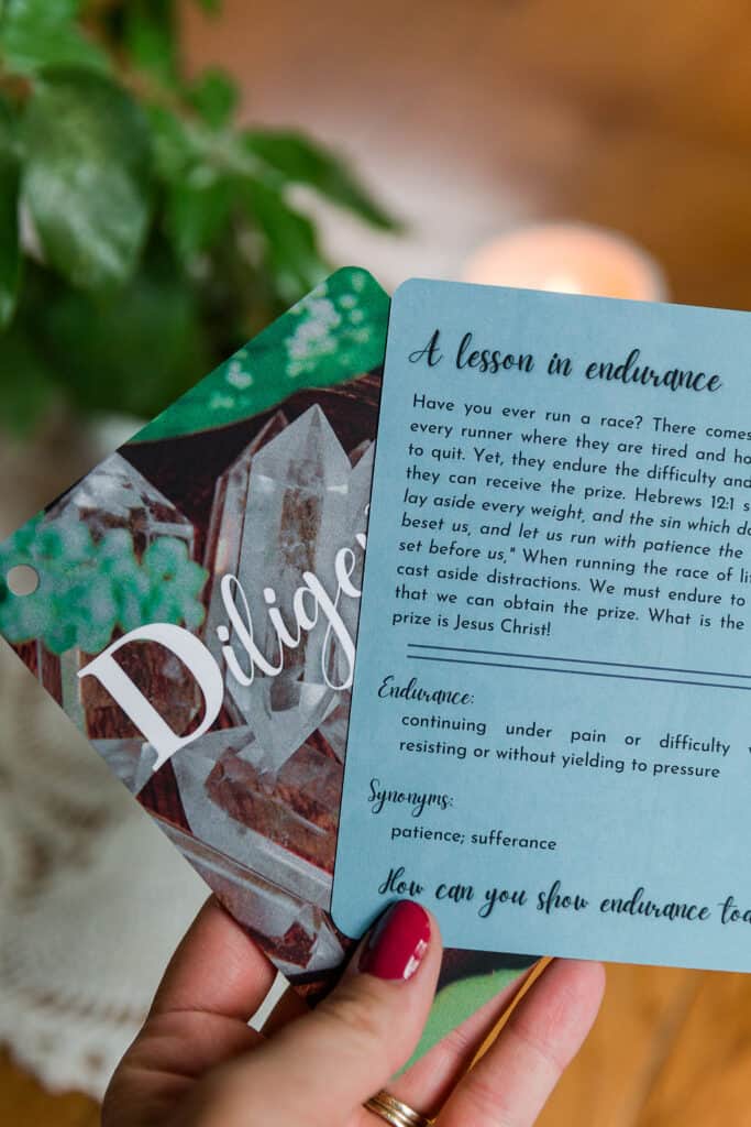 Two "Children of Character Cards" being held in a woman's hand with fingernails painted red. It shows the front and back of the cards to display their design. In the background is a plant on a table which is out of focus.