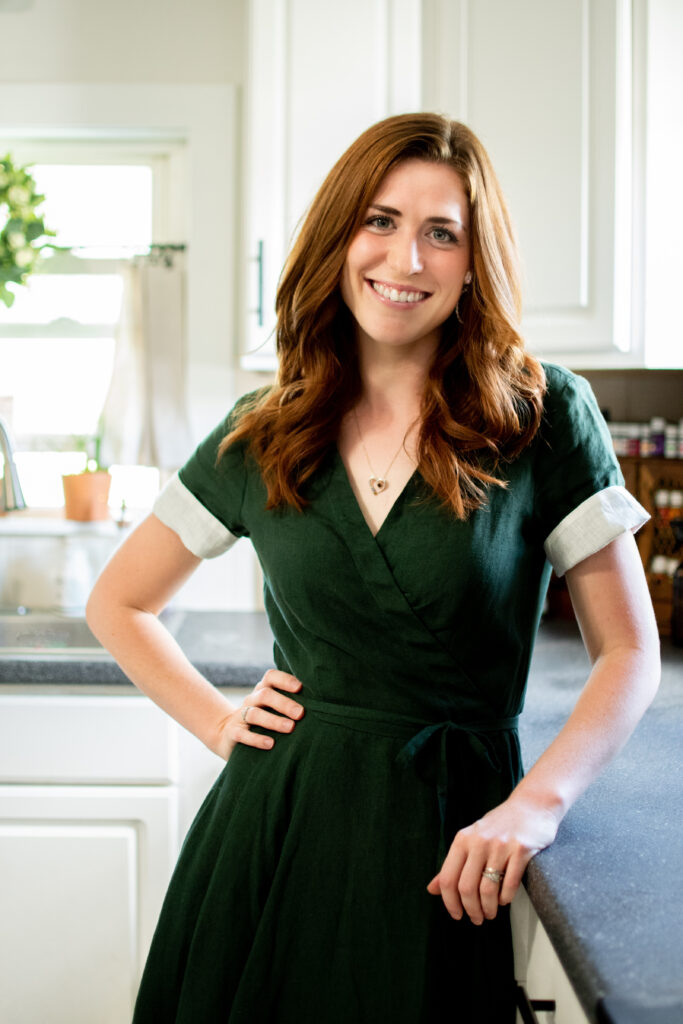 Rebecca Grant is standing in her kitchen leaning against the counter. She is wearing a deep green dress and smiling at the camera.