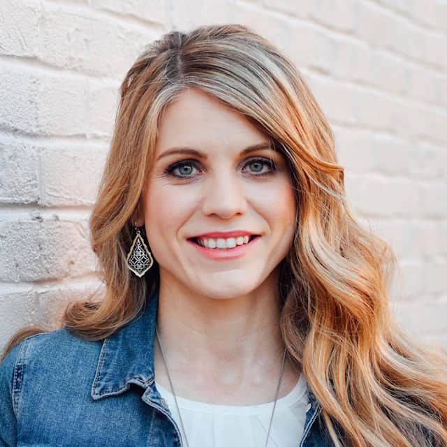 Crystal Paine stands in front of a white brick wall smiling. She is wearing a jean jacket and a white blouse.
