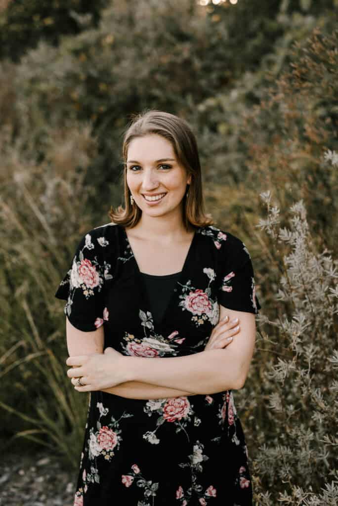 Jami Balmet standing in a black floral dress in front of a field of wild flowers. She is smiling and looking at the camera with her arms crossed in front of her.