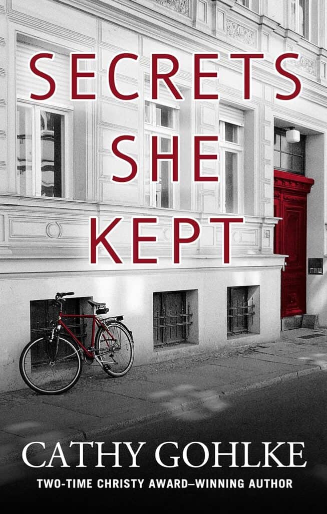 Book cover of "Secrets She Kept" by Cathy Gohlke. It shows a white building with a bicycle leaning against it. There is a bright red door in the background.