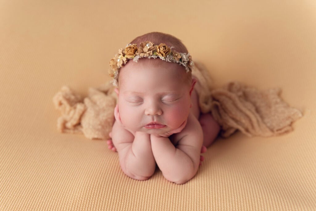 Newborn baby photo. The baby is laying with her hands under her chin. She has a golden colored crown and blanket. She is laying on a golden colored backdrop.