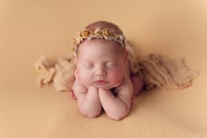 Newborn baby photo. The baby is laying with her hands under her chin. She has a golden colored crown and blanket. She is laying on a golden colored backdrop.