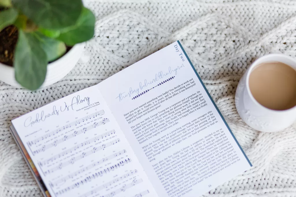 Great Hymns for Growing Hearts Interior Image showing the music to the hymn along side the story behind the hymn. The book sits on a table with hot chocolate and a plant.