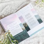 Great Hymns for Growing Hearts book laying open on a table beside a coffee and a bunch of flowers. Interior page shows a hymn title on the left and the words on the right. There is a image of woods and the setting sun in the background.
