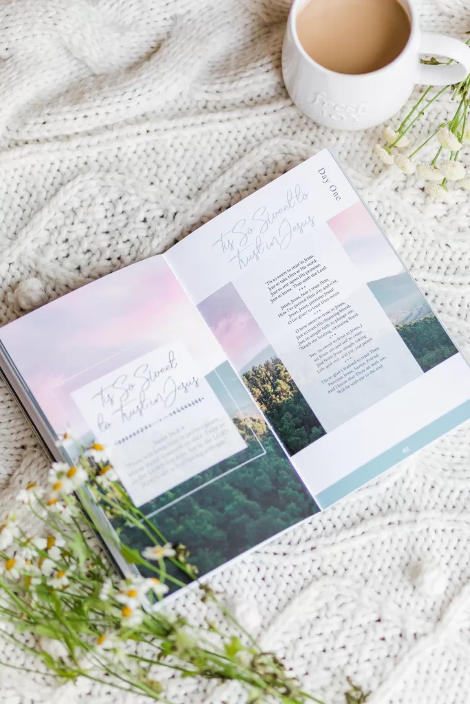 Great Hymns for Growing Hearts book laying open on a table beside a coffee and a bunch of flowers. Interior page shows a hymn title on the left and the words on the right. There is a image of woods and the setting sun in the background.