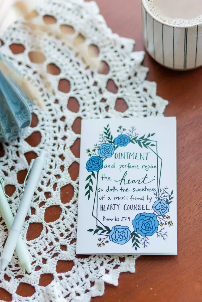 A Friendship card laying on a wooden table with a lace cover. The card reads "Ointment and perfume rejoice the heart: so doth the sweetness of a man's friend by hearty counsel." Proverbs 27:9