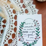 A Friendship Card laying on a wooden table with a lace covering. The card reads "And let us consider one another to provoke unto love and good works:" Hebrews 10:25. The text is wrapped in flowers and leaves