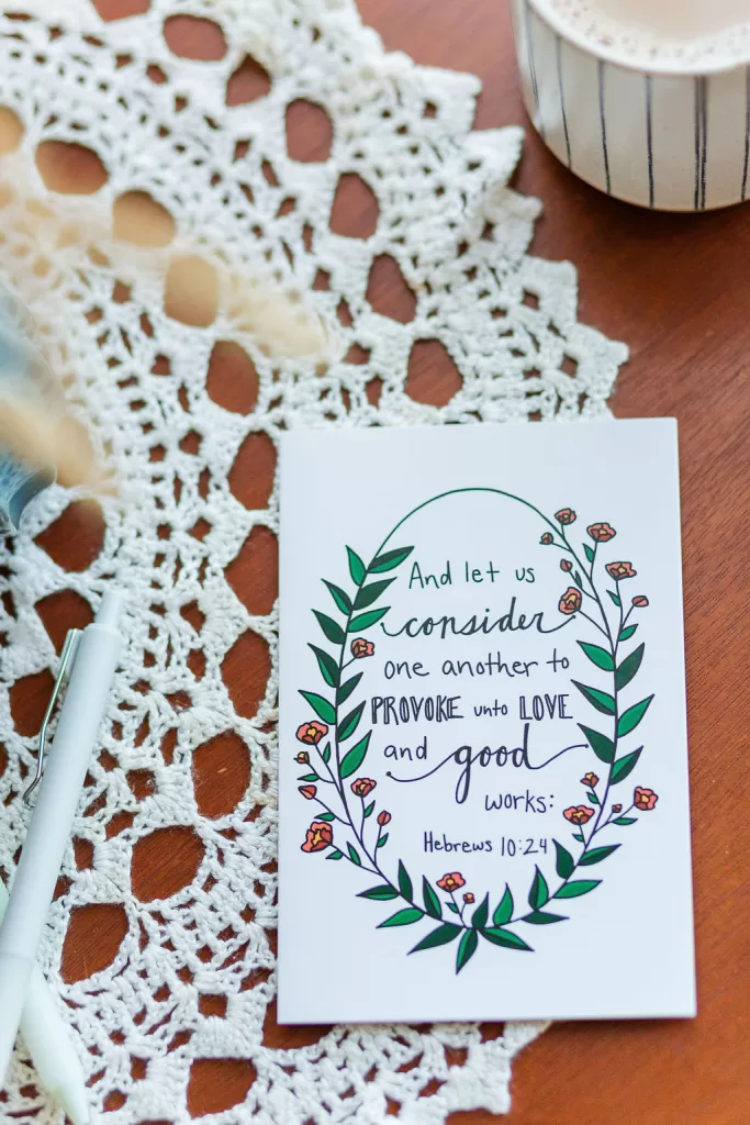 A Friendship Card laying on a wooden table with a lace covering. The card reads "And let us consider one another to provoke unto love and good works:" Hebrews 10:25. The text is wrapped in flowers and leaves
