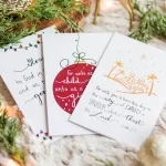 3 of the Christmas cards flayed out on display surrounded by bulbs and greenery. One depicts Bethlehem, one has a bulb, and one as some text that isn't in view.