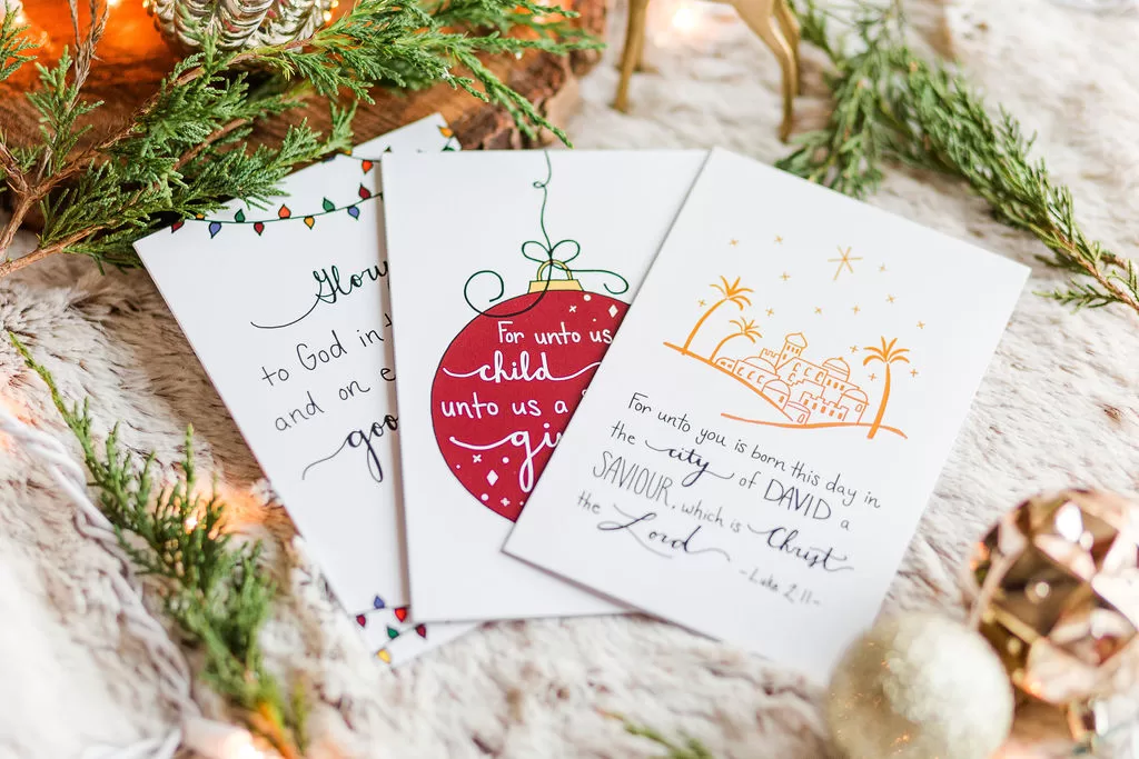 3 of the Christmas cards flayed out on display surrounded by bulbs and greenery. One depicts Bethlehem, one has a bulb, and one as some text that isn't in view.