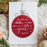 A single card laying on fir and surrounded by greenery. It has a red Christmas bulb and says "For unto us a child is born, unto us a Son is given" Isaiah 9:6