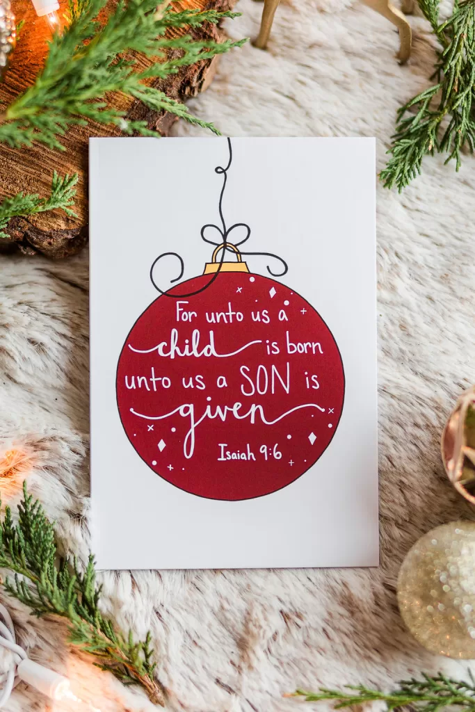A single card laying on fir and surrounded by greenery. It has a red Christmas bulb and says "For unto us a child is born, unto us a Son is given" Isaiah 9:6