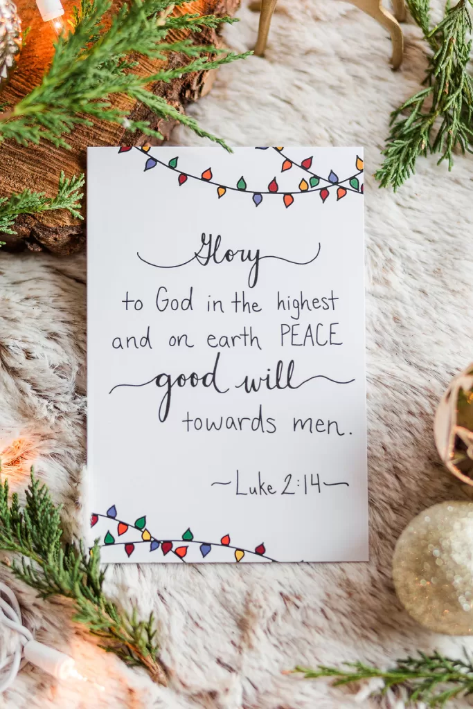 A single Christmas card laying on white fir and surrounded by greenery. It reads "Glory to God in the highest, and on earth Peace, good will towards men." Luke 2:14