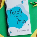 The "Teach Me to Pray Prayer Journal for Kids" laying on two sheets of yellow and green construction paper with colored pencils next to it.