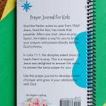 The Teach Me To Pray Prayer Journal laying on a piece of green and yellow construction paper on top of a table. There are colored pencils beside it. It shows the back of the journal with the description.