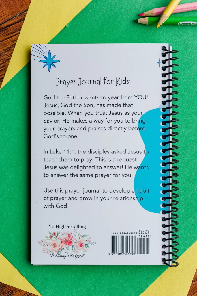 The Teach Me To Pray Prayer Journal laying on a piece of green and yellow construction paper on top of a table. There are colored pencils beside it. It shows the back of the journal with the description.