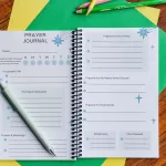 The Teach Me To Pray Prayer Journal laid open to show the prayer request and prompt areas. It is on top of colored construction paper on a wooden table. There are colored pencils beside it.
