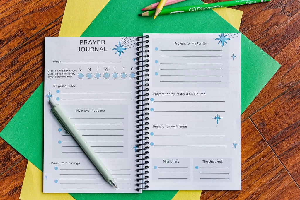 The Teach Me To Pray Prayer Journal laid open to show the prayer request and prompt areas. It is on top of colored construction paper on a wooden table. There are colored pencils beside it.